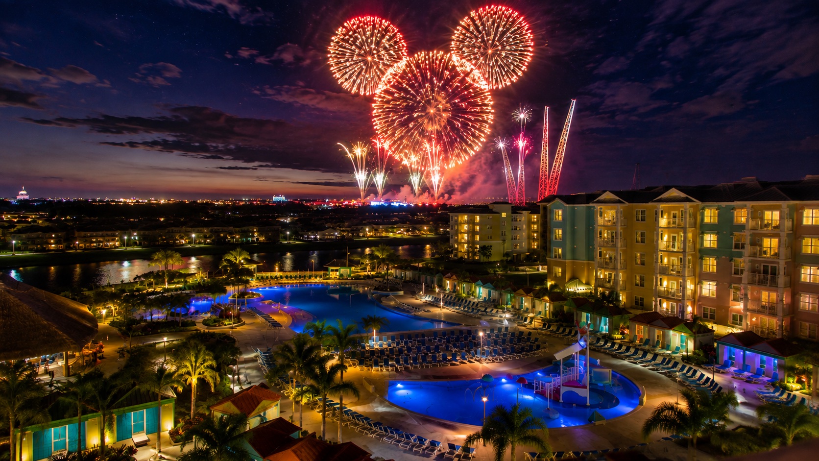 Night view over the resort at Sunset Walk from an upper floor: lit lagoon and zero-entry pools ringed by palms and loungers, resort buildings glowing at right, and a fireworks burst over the treeline in the distance.